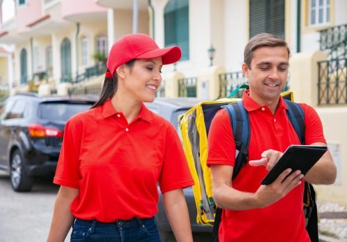 Happy man in red cap showing address to colleague Happy man in red cap showing address to colleague. Smiling couriers working together and delivering orders on foot. Houses and cars on background. Delivery service and online shopping concept